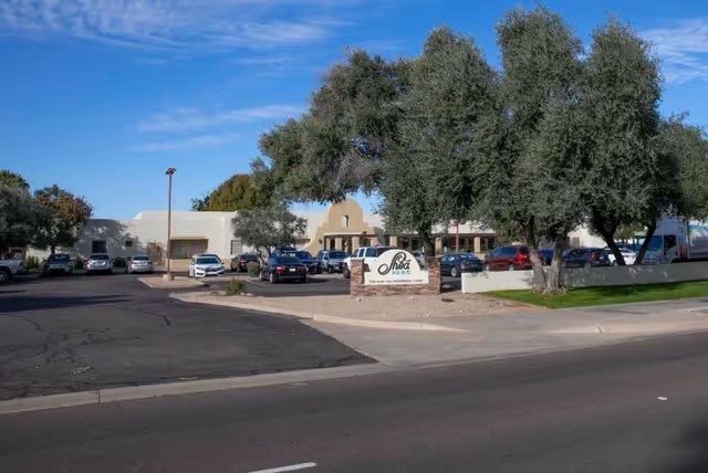 Exterior view of Shea Post Acute Rehabilitation Center showing a single-story building with a parking lot in front, several cars parked, trees around the area, and a sign with the facility's name near the entrance.