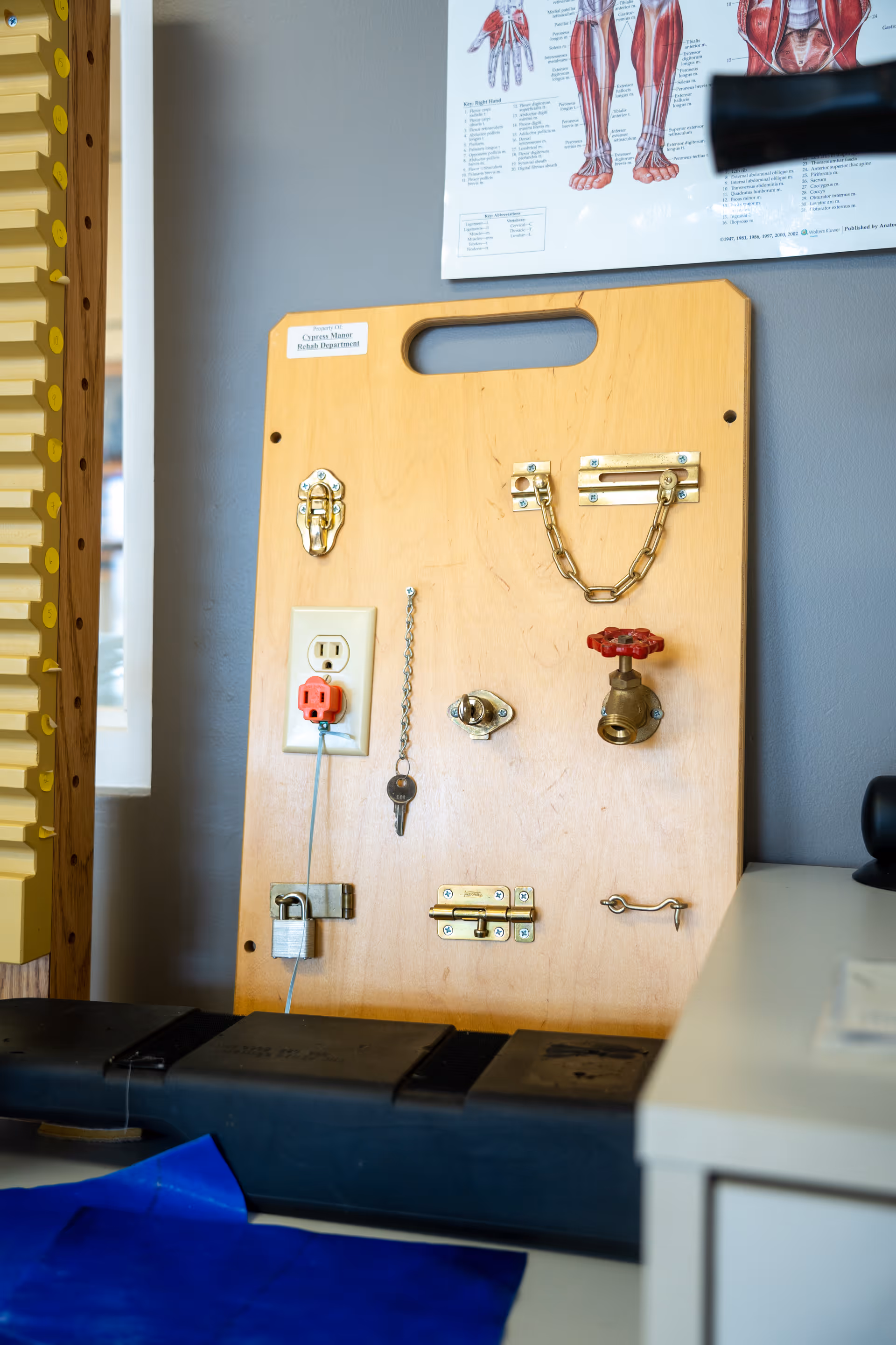 A wooden therapy/activity board on a table with various latches, locks, a faucet valve, electrical outlet and hanging key in a clinic room.