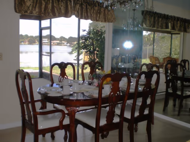 A dining room with a wooden dining table set for six people with white plates, cups, and utensils. The room has large windows with a view of a lake and greenery outside. There is a decorative curtain above the windows and a chandelier hanging from the ceiling. A mirror on the right side reflects part of the room and the outdoor scenery.