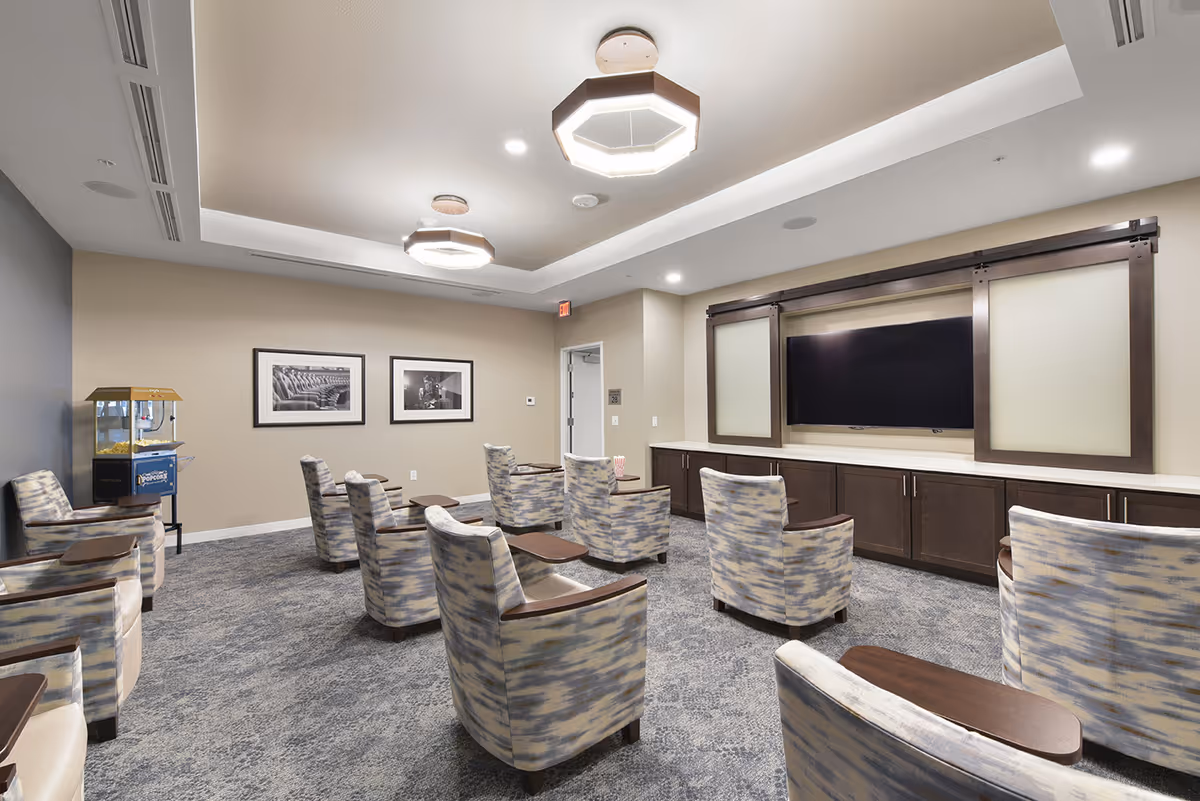 A senior living community media room with upholstered chairs arranged facing a large wall-mounted TV and a popcorn machine at the side.
