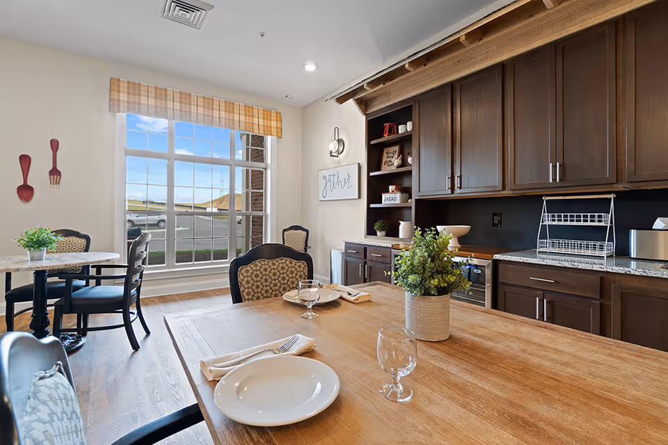 A bright dining area with a wooden table set with plates, glasses, and napkins. The room features dark wooden cabinets, a countertop with a dish rack, and a large window with a plaid valance letting in natural light. There are additional small tables and chairs near the window, and decorative wall art including oversized wooden spoon and fork and a sign that says 'gather'.