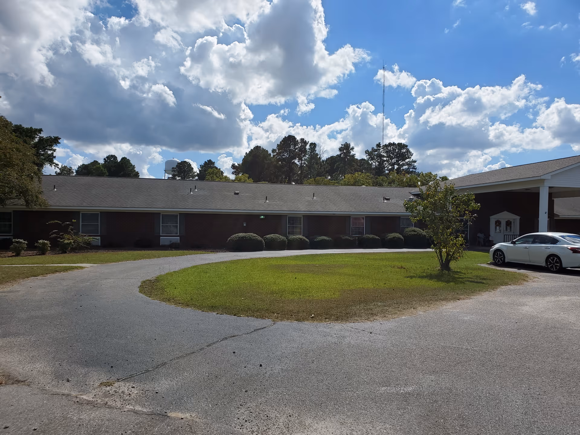 Single-story brick senior living building with a circular driveway and grassy center under a partly cloudy sky.