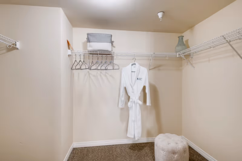Walk-in closet with wire shelving, multiple hangers, a white robe hanging in the center, a storage basket on the shelf and a tufted ottoman on the floor.