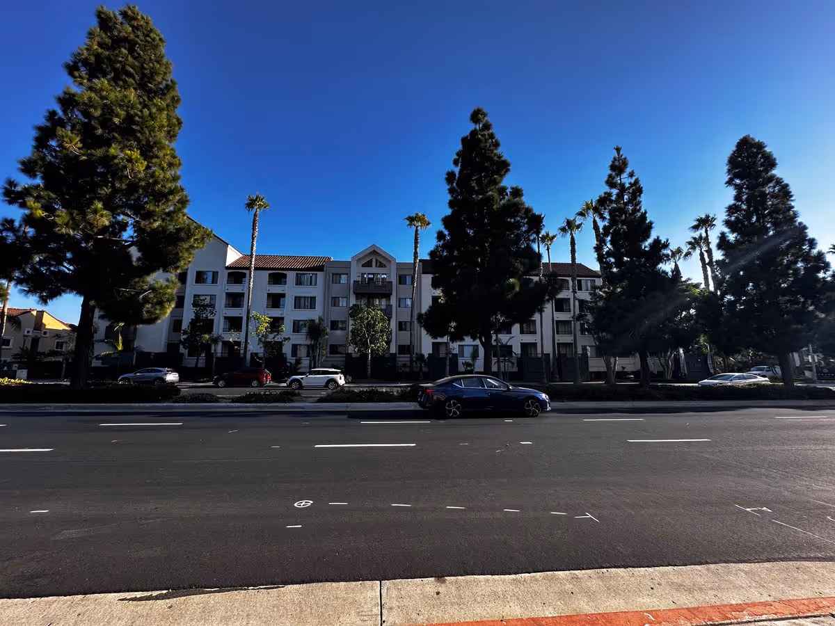 Front exterior of a multi-story white residential building with tall trees and parked cars along a street under a clear blue sky.