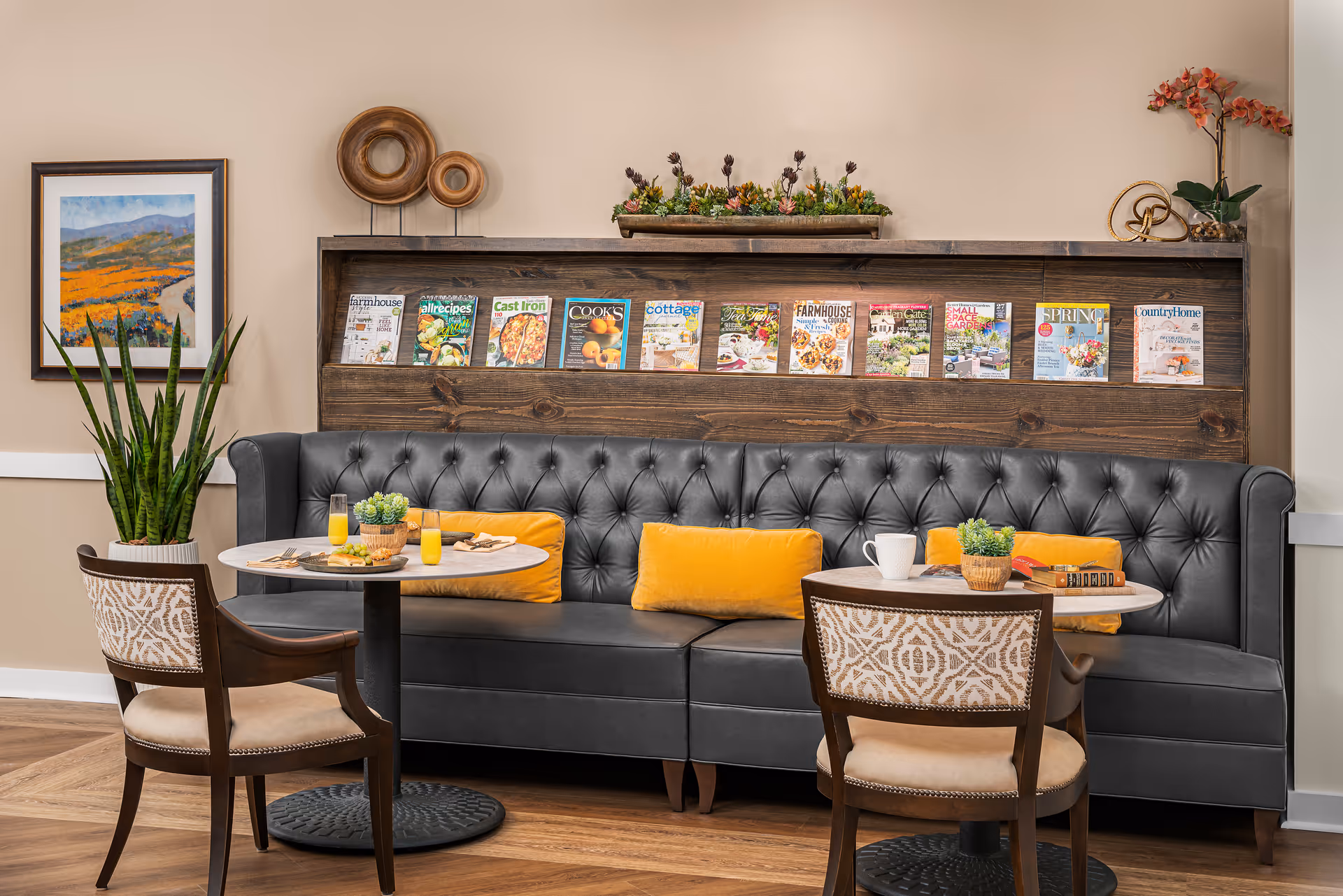 A cozy seating area with a dark gray tufted leather bench adorned with three yellow cushions. In front of the bench are two round tables, each with a wooden chair featuring a patterned backrest. On the tables are breakfast items including glasses of orange juice, a white coffee mug, and plates with food. Behind the bench is a wooden shelf displaying various magazines and decorative plants. The wall is beige with framed artwork and additional decorative items.