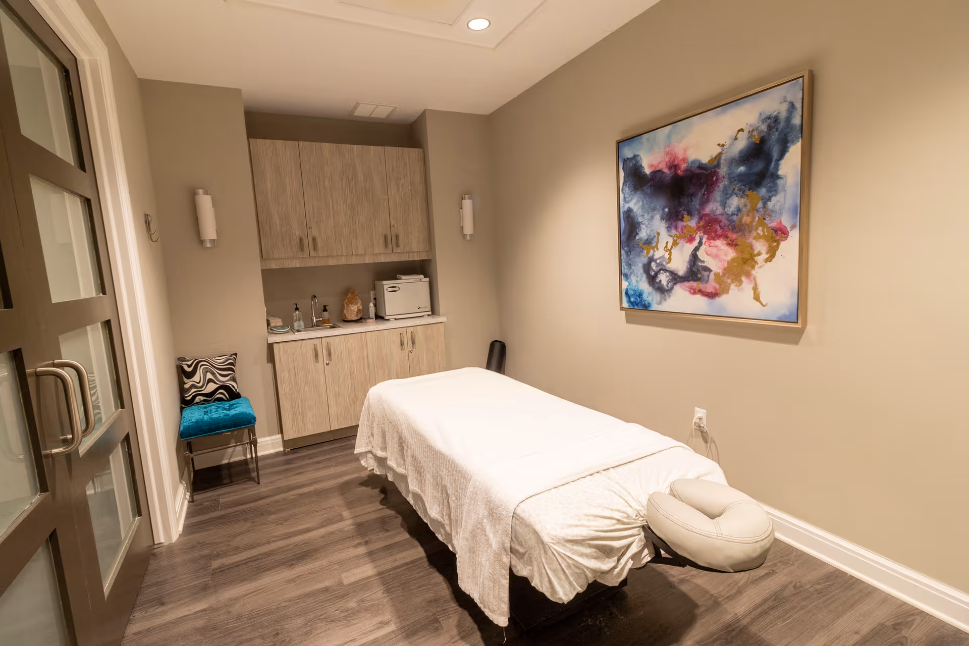 A serene massage or therapy room with a massage table covered in a white sheet, a colorful abstract painting on the wall, wooden cabinets with various items on the countertop, a teal chair with a black and white cushion, and soft lighting.