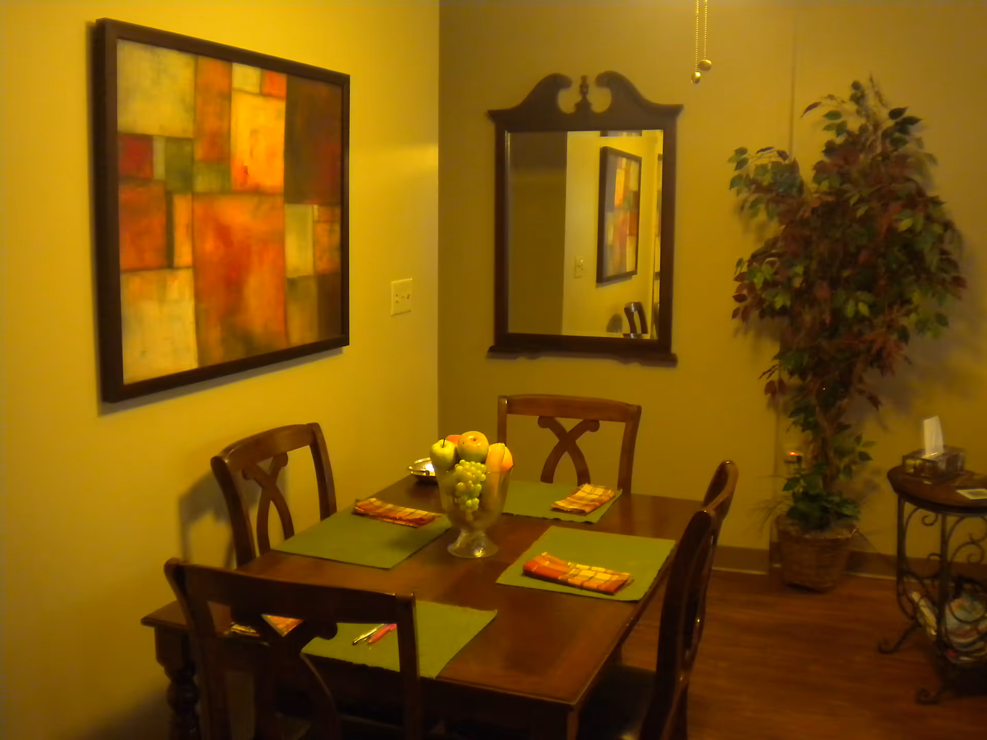 A dining room with a wooden table set for four, featuring green placemats and folded napkins. A glass bowl filled with artificial fruit is centered on the table. On the wall, there is a large abstract painting with warm colors and a decorative mirror reflecting part of the room. A tall potted plant stands in the corner next to a small side table with a tissue box and other items.