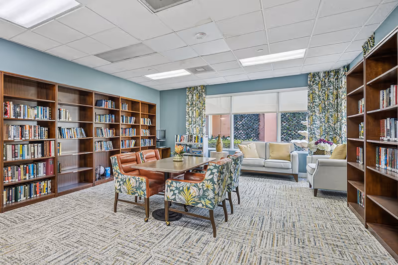 A bright and spacious library room with large wooden bookshelves filled with books along the walls. In the center, there is a rectangular table surrounded by six chairs, four with floral patterned upholstery and two with brown leather upholstery. At the back, there is a seating area with a white sofa and an armchair near large windows with floral curtains, allowing natural light to fill the room.