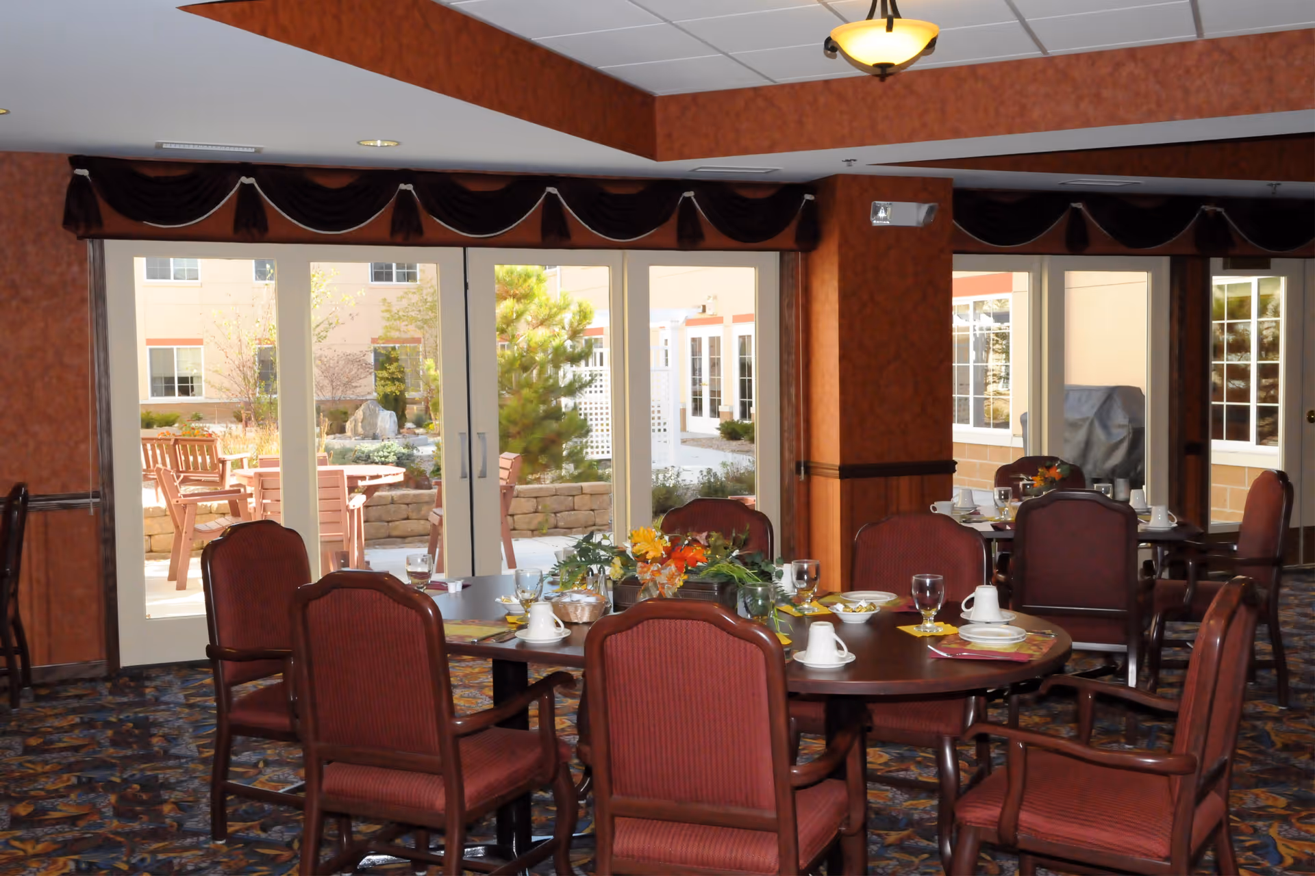 Dining room with round wooden tables and red upholstered chairs set with cups, plates, and glasses. Large windows and glass doors provide a view of an outdoor patio area with wooden benches and greenery.