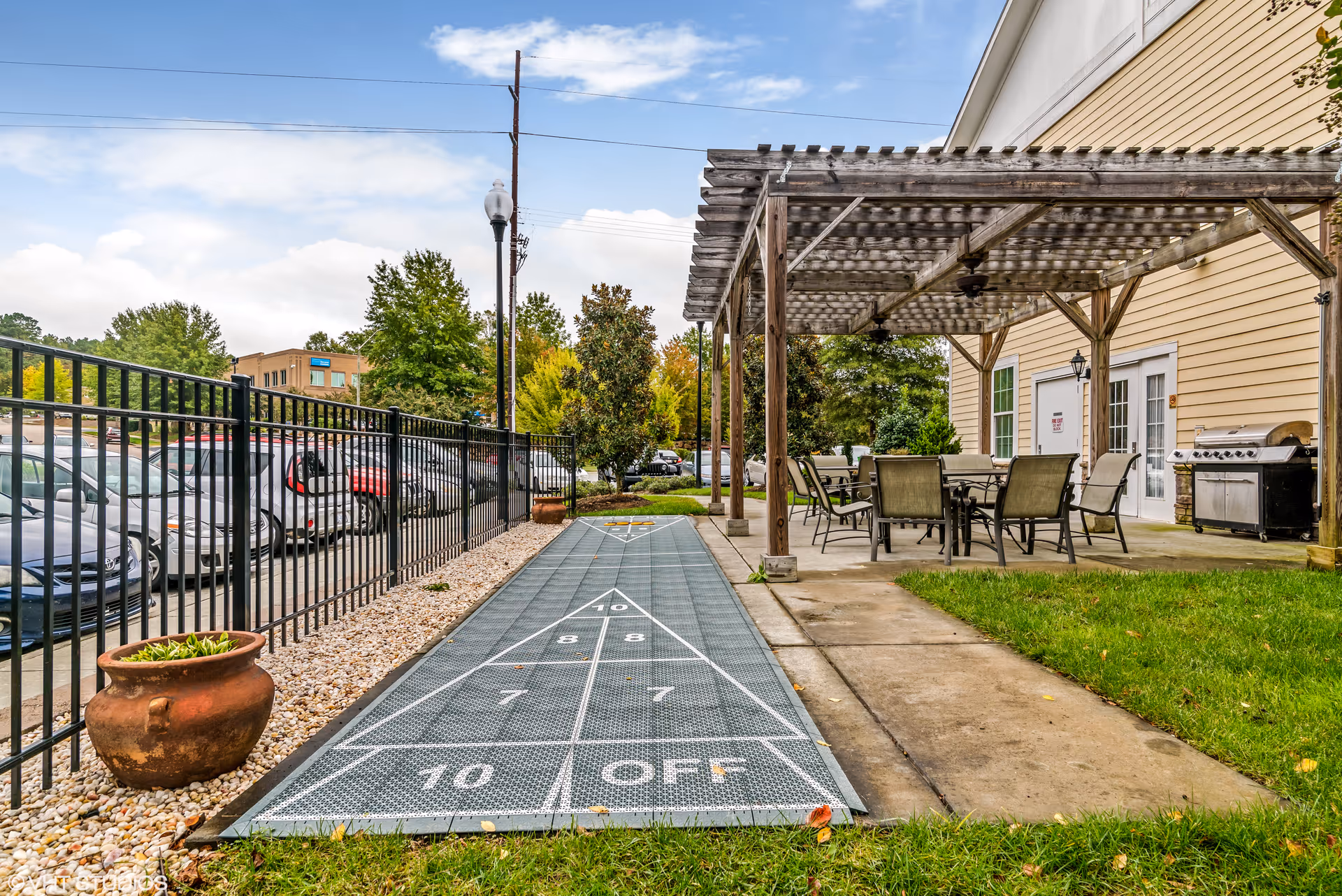 Outdoor patio area with a shuffleboard court, pergola-covered dining table and chairs, a grill, and an adjacent parking lot.