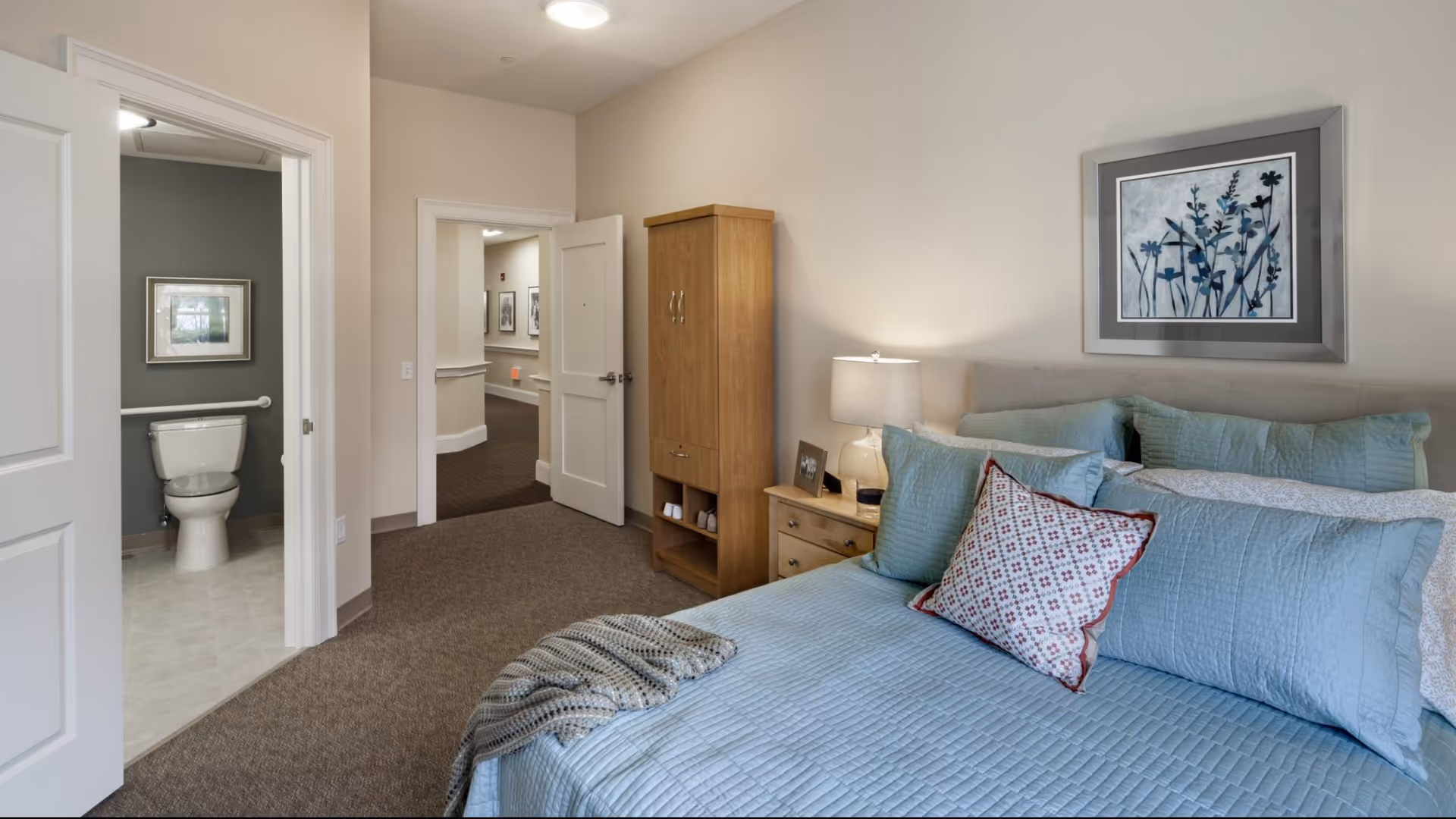 A bedroom in a senior living facility with a neatly made bed featuring blue bedding and multiple pillows, a wooden nightstand with a lamp and picture frame, a wooden cabinet, and a framed floral artwork on the wall. The door to an adjacent bathroom with a toilet is open, and a hallway is visible beyond the bedroom door.