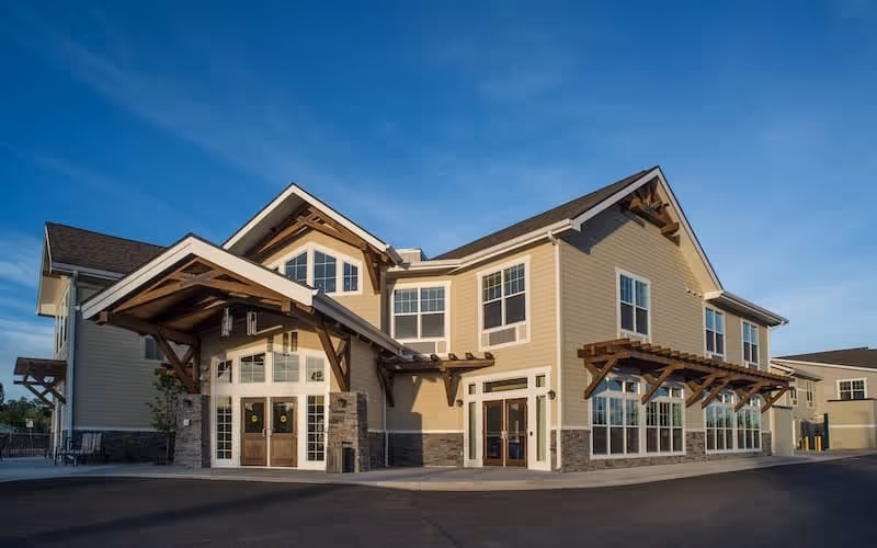 Two-story beige senior living facility with a covered wooden entrance, large windows, and stone accents under a blue sky.