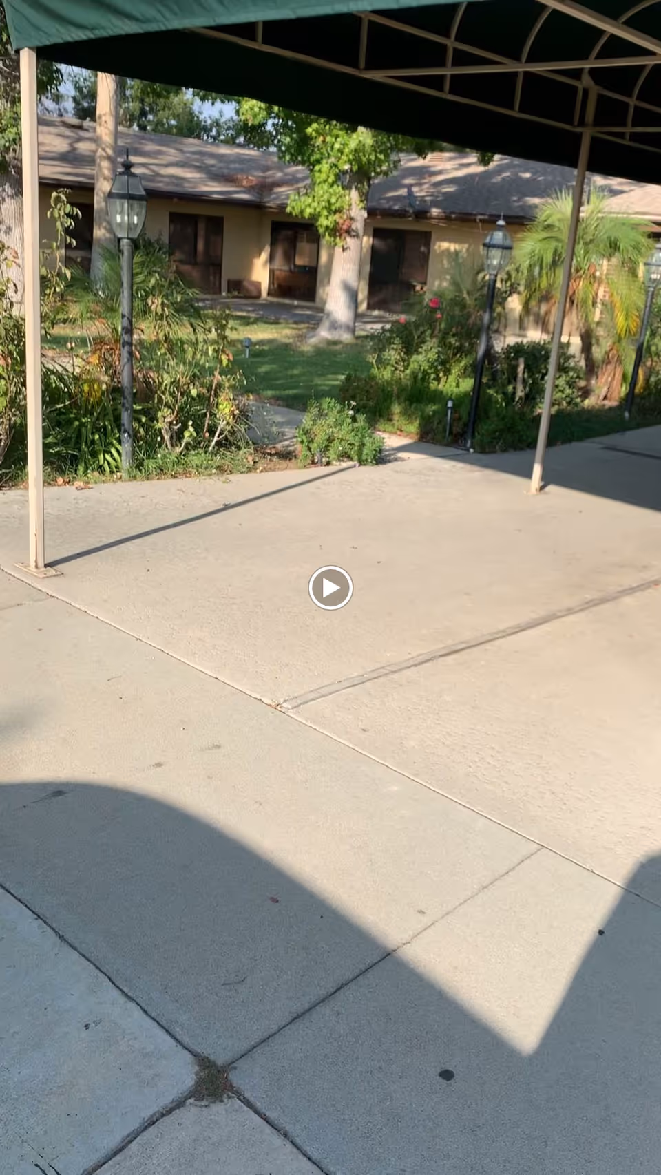Outdoor view of a garden area at Santa Anita Assisted Living, showing a concrete walkway with a green canopy overhead, lamp posts, various plants, and a building with windows in the background.