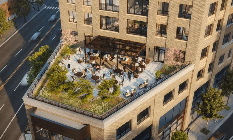 A rooftop outdoor patio area on a multi-story brick building with tables, chairs, and people socializing. The patio is surrounded by greenery and fencing, with a pergola providing shade over some seating. The building is located next to a street with trees and crosswalks visible.