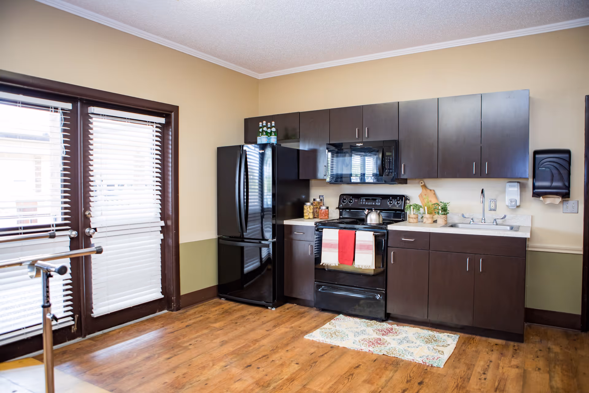 A kitchen area with dark brown cabinets, a black refrigerator, black stove with a red and white towel hanging on the oven door, a microwave above the stove, a sink with a soap dispenser, and a paper towel dispenser mounted on the wall. There are glass jars and small potted plants on the countertop. The floor is wooden, and there is a colorful rug in front of the stove. To the left, there are glass doors with white blinds.