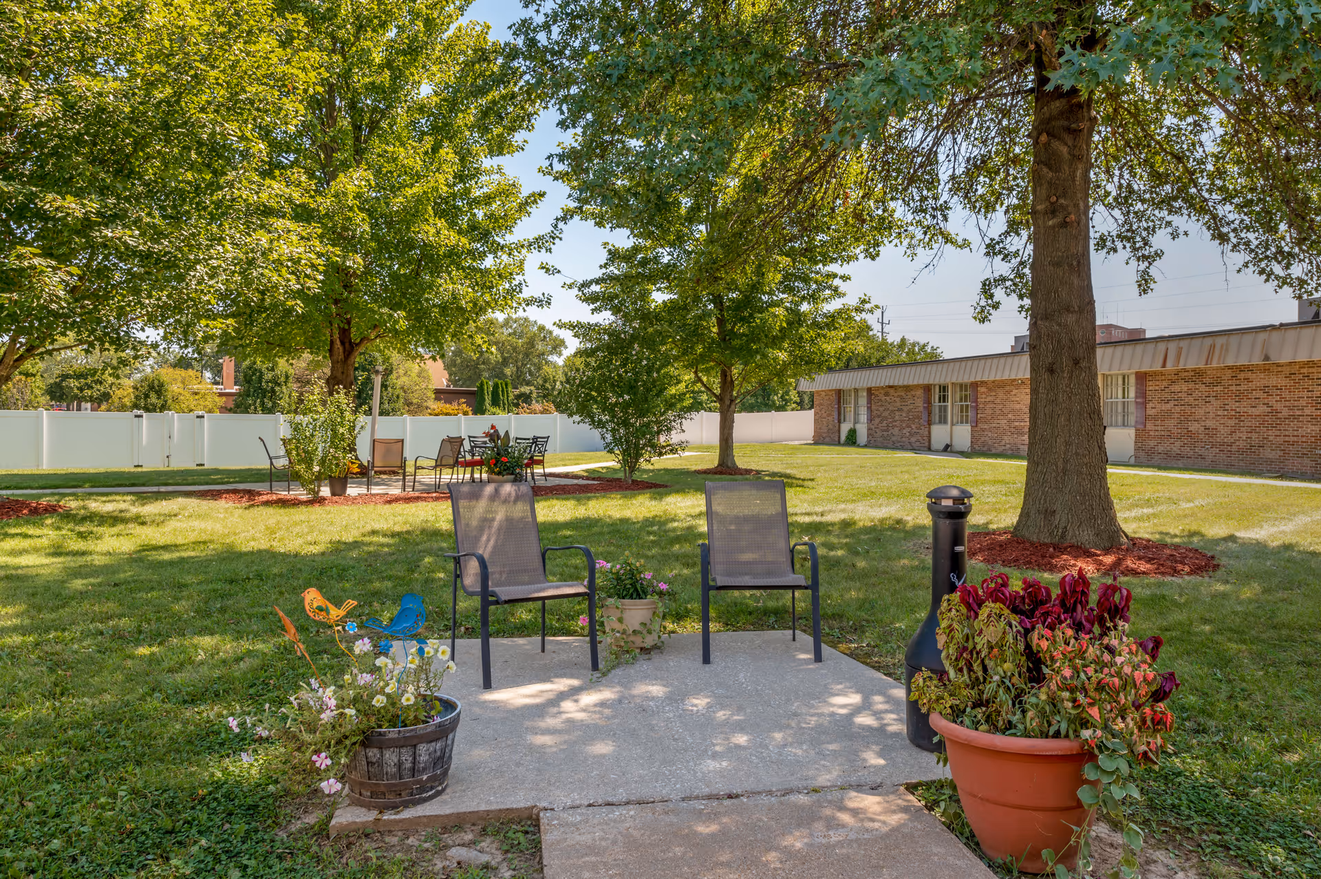 Outdoor seating area in a garden with two chairs on a concrete slab, surrounded by potted plants and trees. In the background, there is a brick building and additional seating with tables and chairs on the grass under trees.