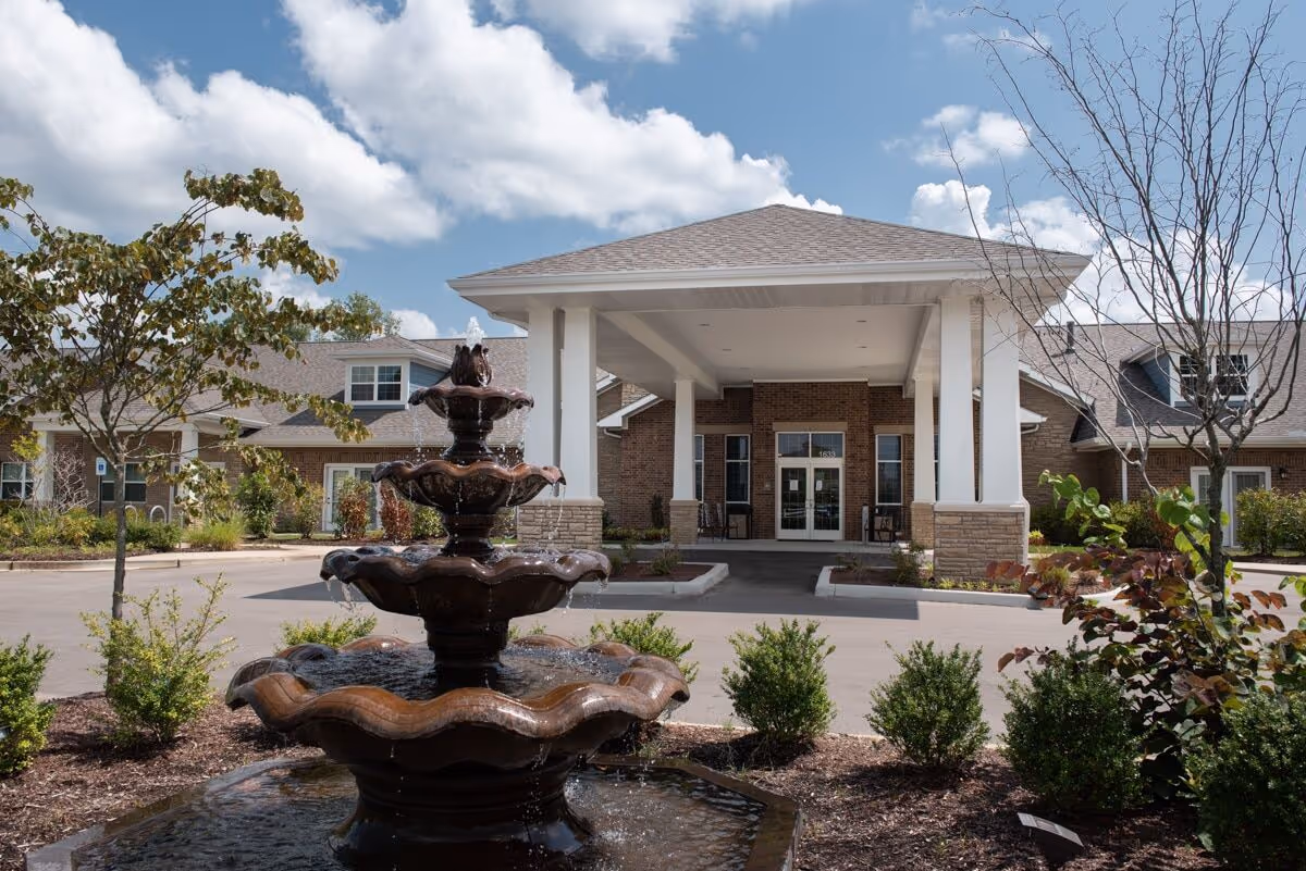 Front exterior view of a senior living facility with a covered entrance supported by white columns, a multi-tiered water fountain in the foreground, landscaped bushes and trees, and a partly cloudy sky.
