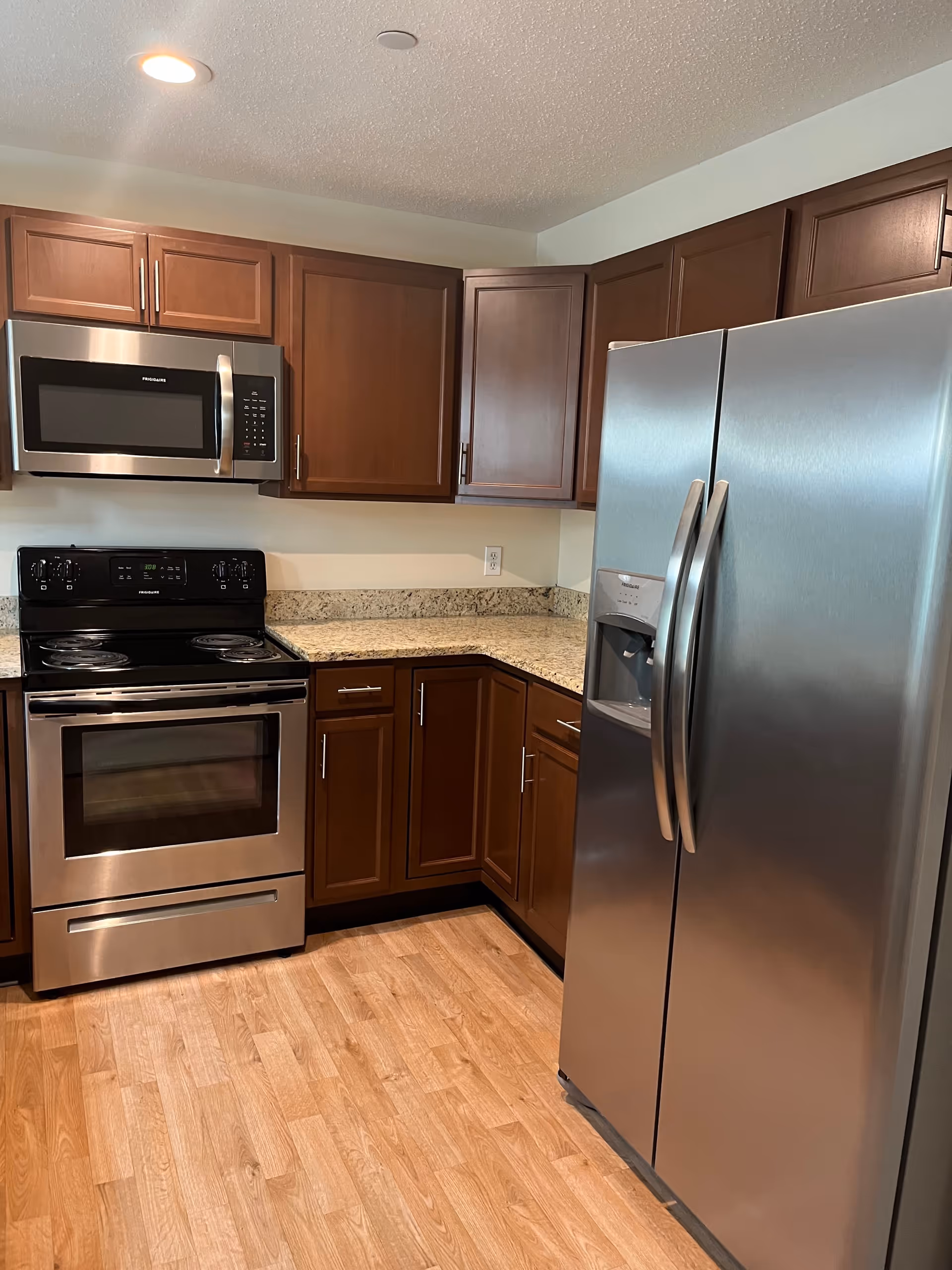 Modern kitchen with stainless steel appliances including a refrigerator, microwave, and stove. The kitchen has wooden cabinets and granite countertops with a wood-patterned floor.