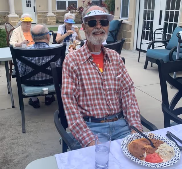 An elderly man wearing sunglasses, a white visor, and a red plaid shirt sits at an outdoor table with a plate of food and a glass of water in front of him. In the background, three other elderly individuals are seated at another table, also wearing visors and enjoying a meal outside near a building with large windows and doors.