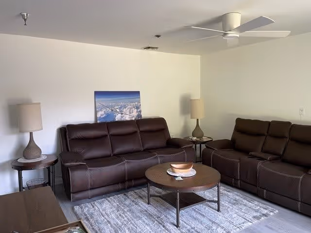 A living room with two dark brown leather sofas facing each other, a round wooden coffee table with a decorative bowl on top, two side tables each with a lamp, a ceiling fan, and a framed picture of clouds on the wall.