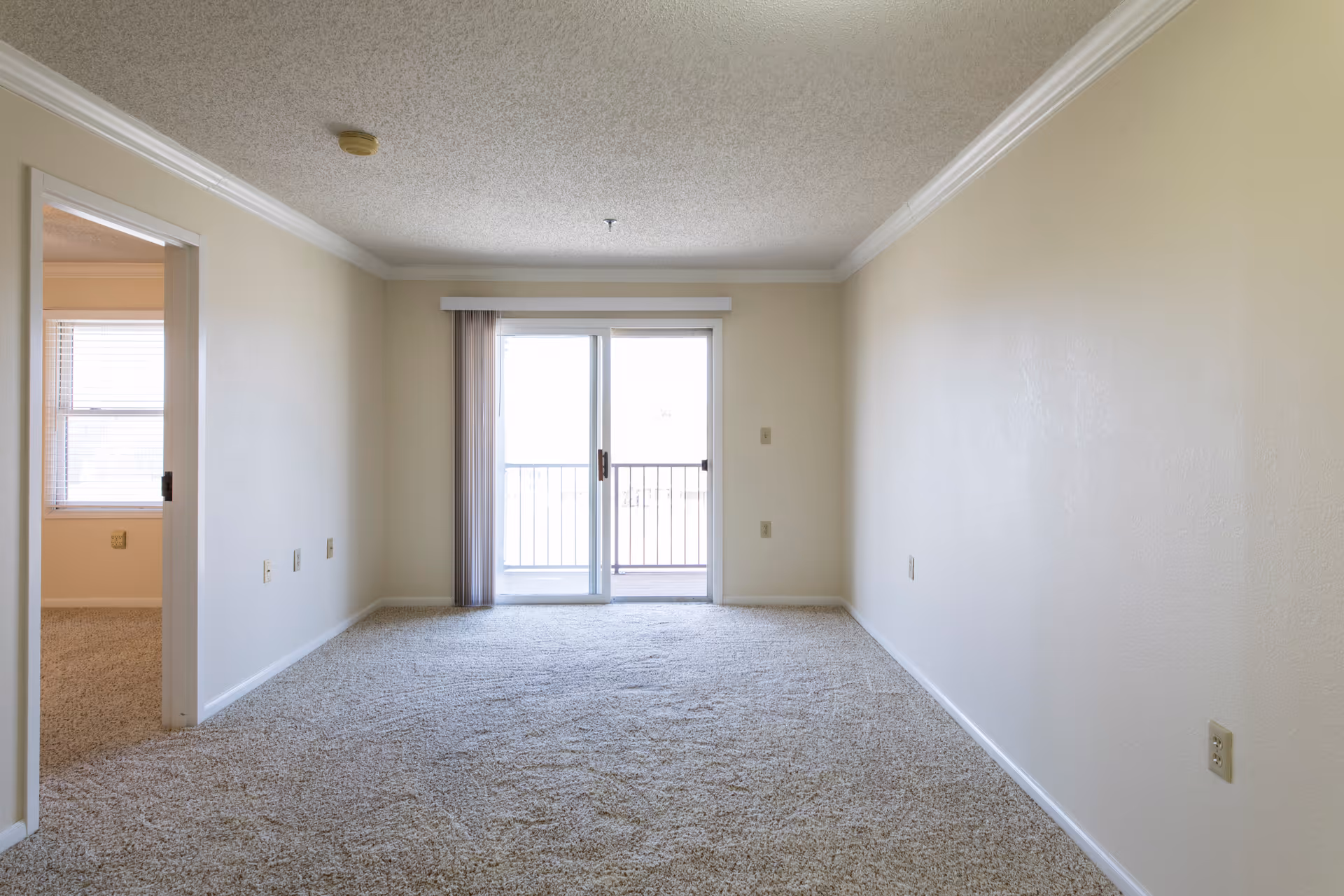 Empty room with beige carpet and cream-colored walls, featuring a sliding glass door leading to a balcony with vertical blinds partially open. To the left, there is an open doorway leading to another room with a window and similar carpeting. The ceiling has a textured finish with crown molding along the edges.