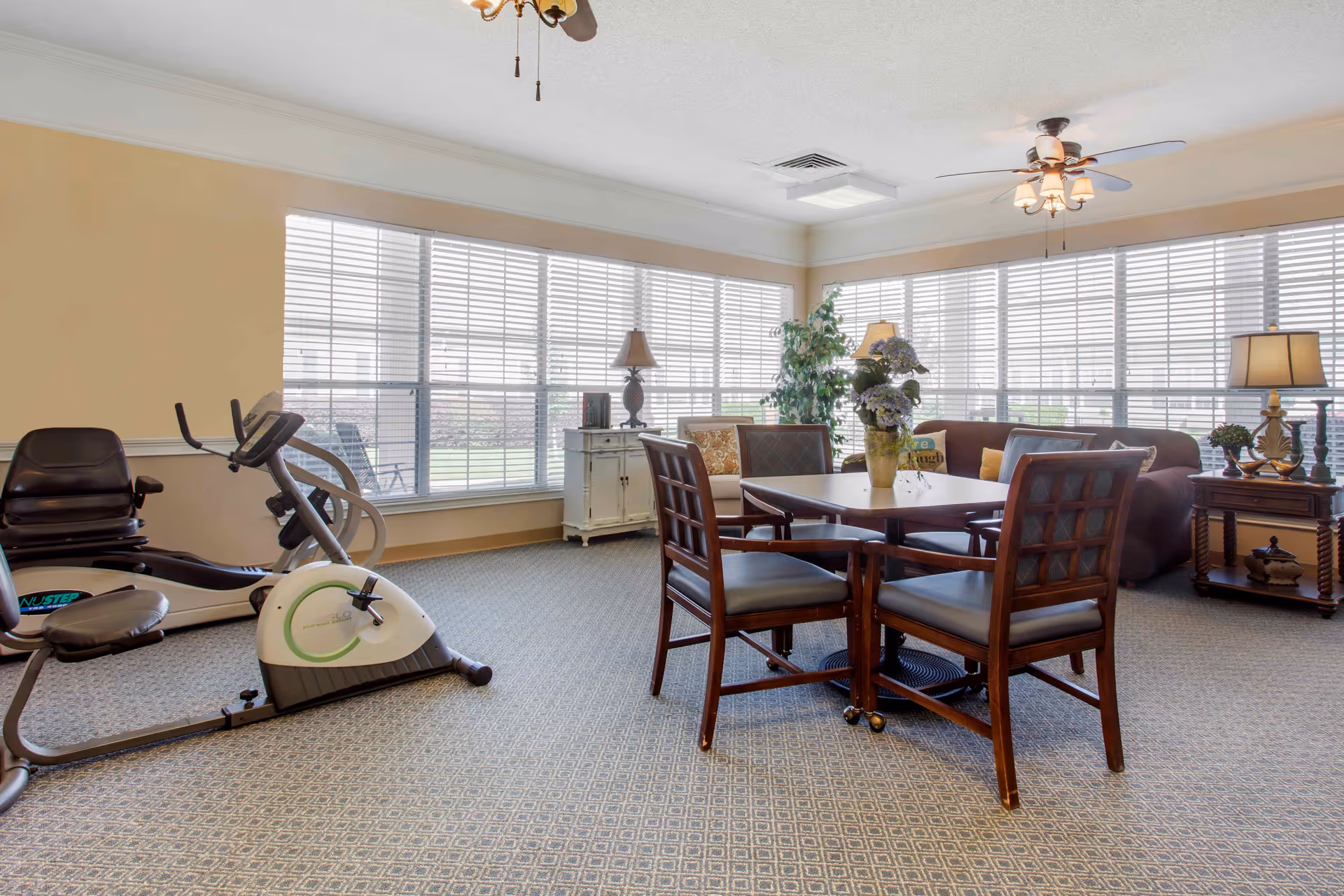 A bright room with large windows covered by white blinds. The room contains exercise equipment including a recumbent bike and an elliptical machine on the left side. In the center, there is a wooden table with four chairs around it, and a vase with flowers on the table. On the right side, there is a brown sofa with cushions, a side table with a lamp, and a potted plant near the windows. The room has a carpeted floor and ceiling fans with lights.