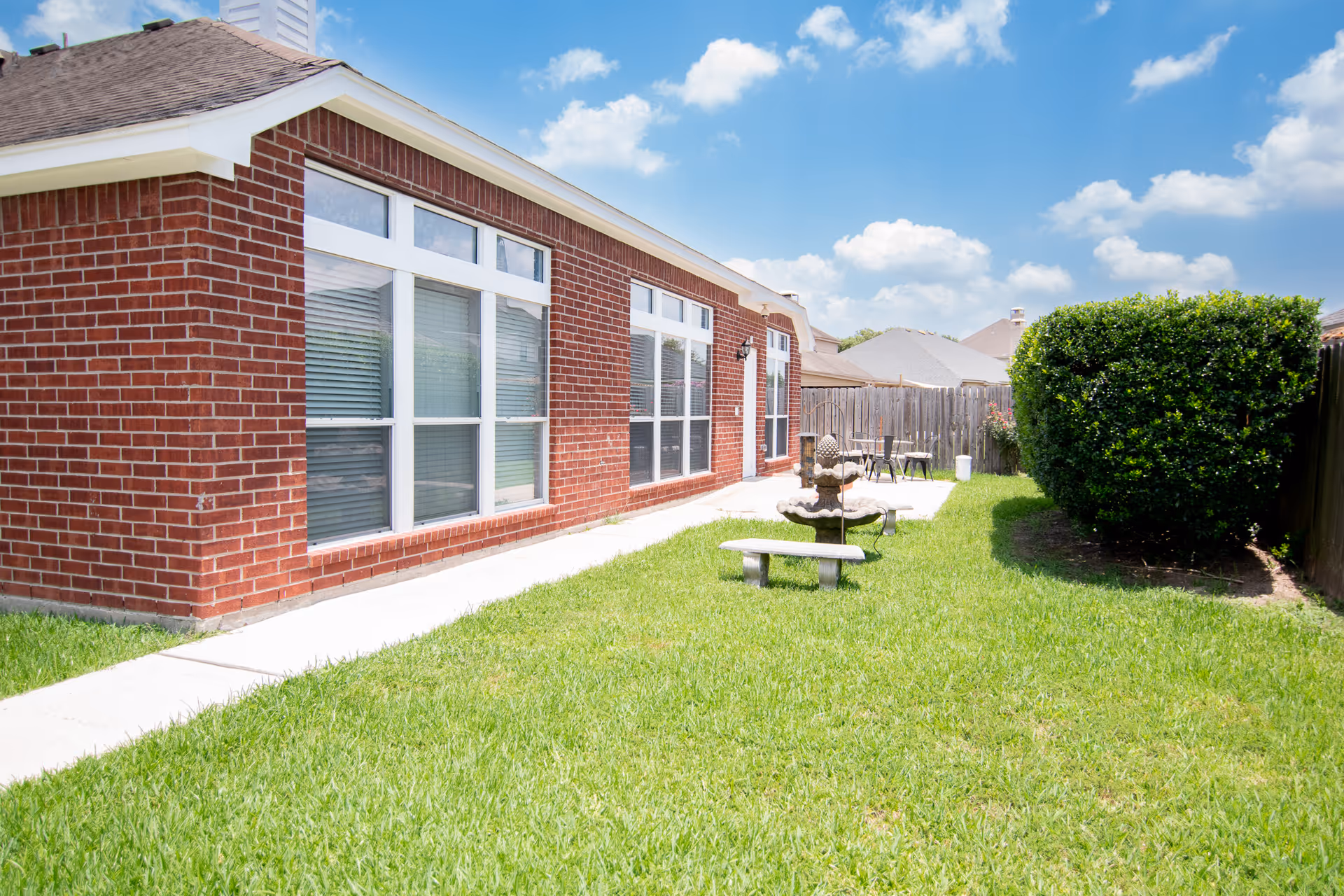Outdoor view of a brick building with large windows and a concrete pathway alongside a green lawn. There is a small stone bench and a decorative fountain in the grass, with a wooden fence and trimmed bushes in the background under a partly cloudy blue sky.