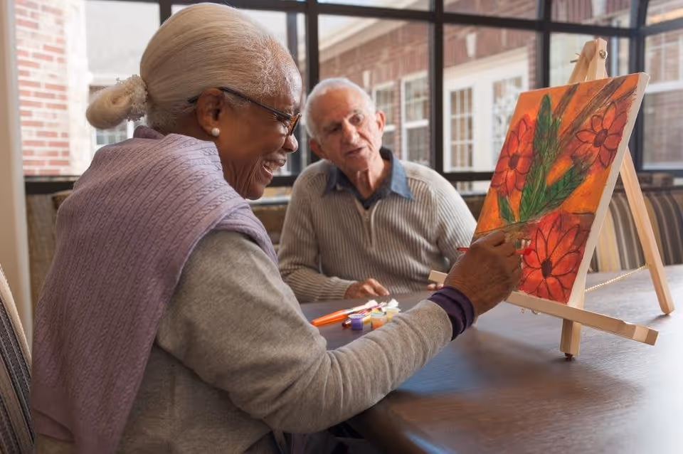 An elderly woman with gray hair tied back and wearing glasses and a lavender shawl is painting a colorful floral picture on a canvas set on an easel. An elderly man with white hair and a striped sweater sits across the table, watching her paint. They are in a bright room with large windows and brick walls.