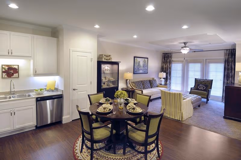 A bright and spacious open-concept living area in a senior living facility featuring a kitchen with white cabinets and stainless steel dishwasher, a round dining table set for four with green cushioned chairs, and a cozy living room with a sofa, armchair, ottoman, and large windows with curtains allowing natural light.