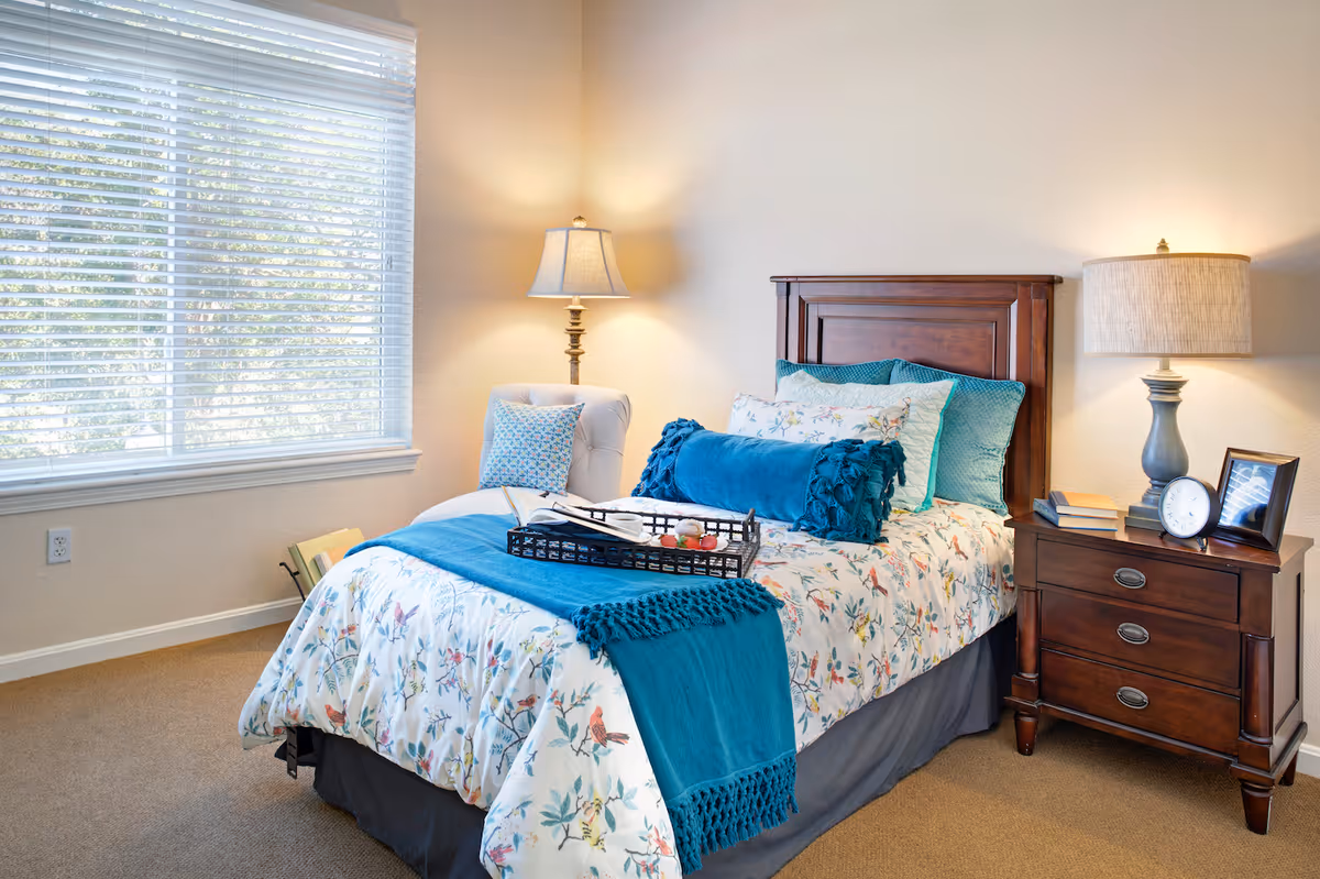 Well-lit bedroom with a single bed dressed in floral bedding and teal accents, a nightstand with a lamp and clock, and a large window.
