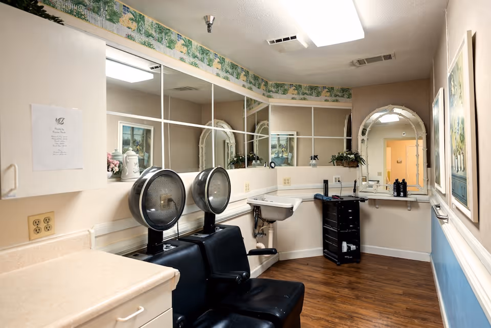 Interior view of a hair salon area within a senior living facility. The room features two black salon chairs with attached hair dryers, a small white sink, a black rolling cart with hair products, a large mirror with a decorative frame, and a wall covered with square mirror tiles. The floor is wooden, and the walls are decorated with floral wallpaper border and framed artwork.