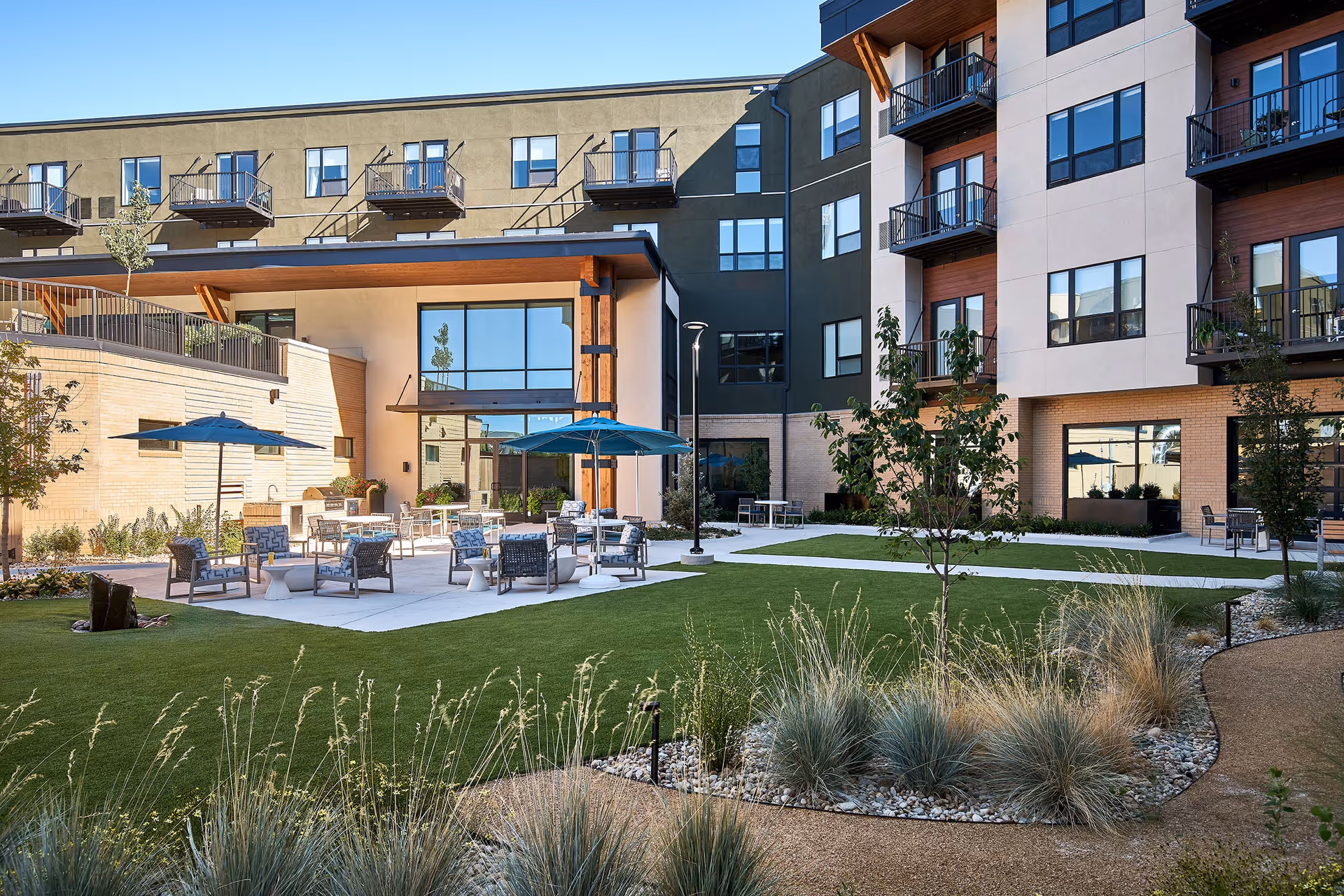 Outdoor courtyard with lawn, patio seating and umbrellas in front of a multi-story senior living building with balconies.