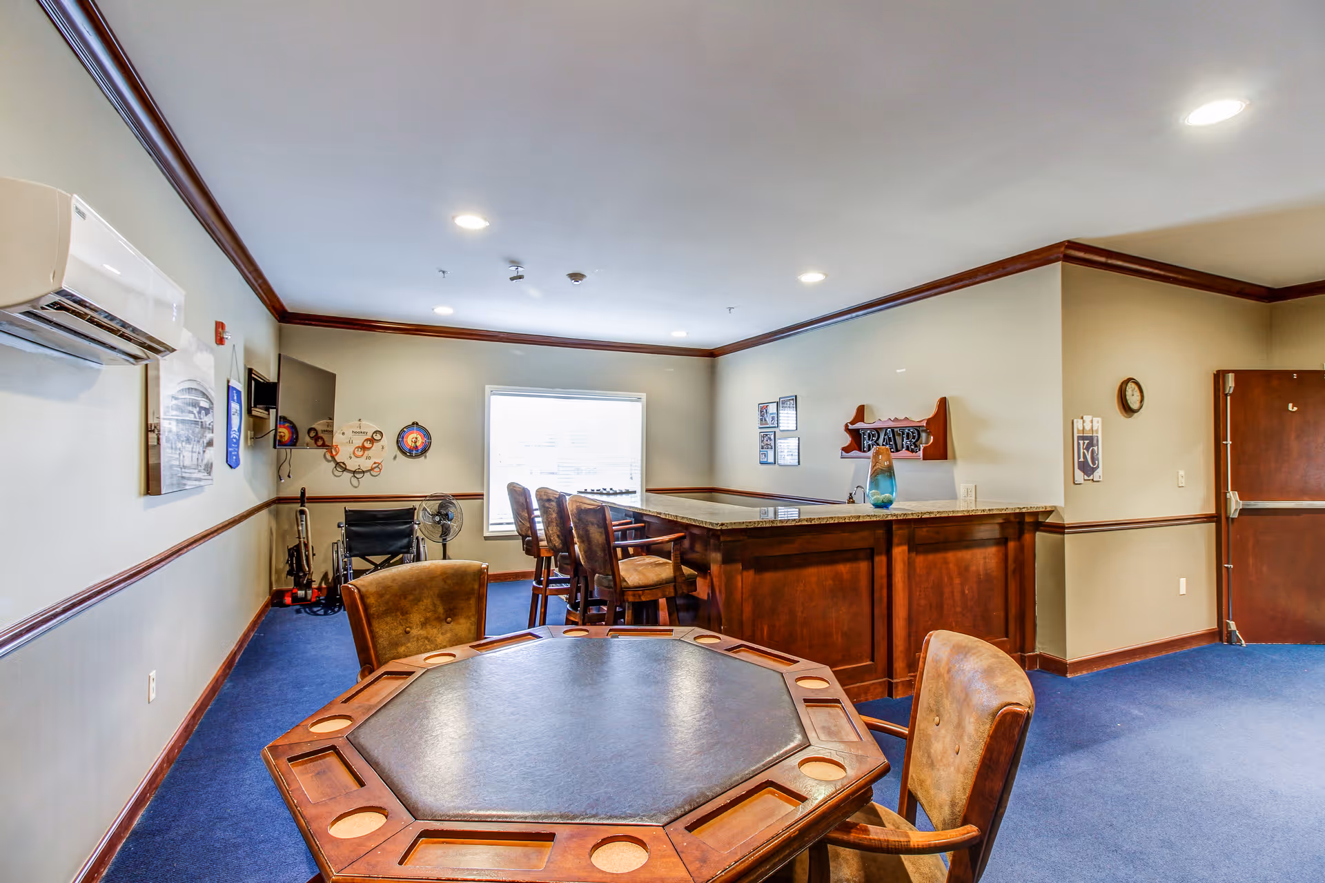 Interior room with a wooden octagonal table and chairs in the foreground, a bar counter with high chairs in the background, blue carpet, beige walls with dark wood trim, and a window letting in natural light.