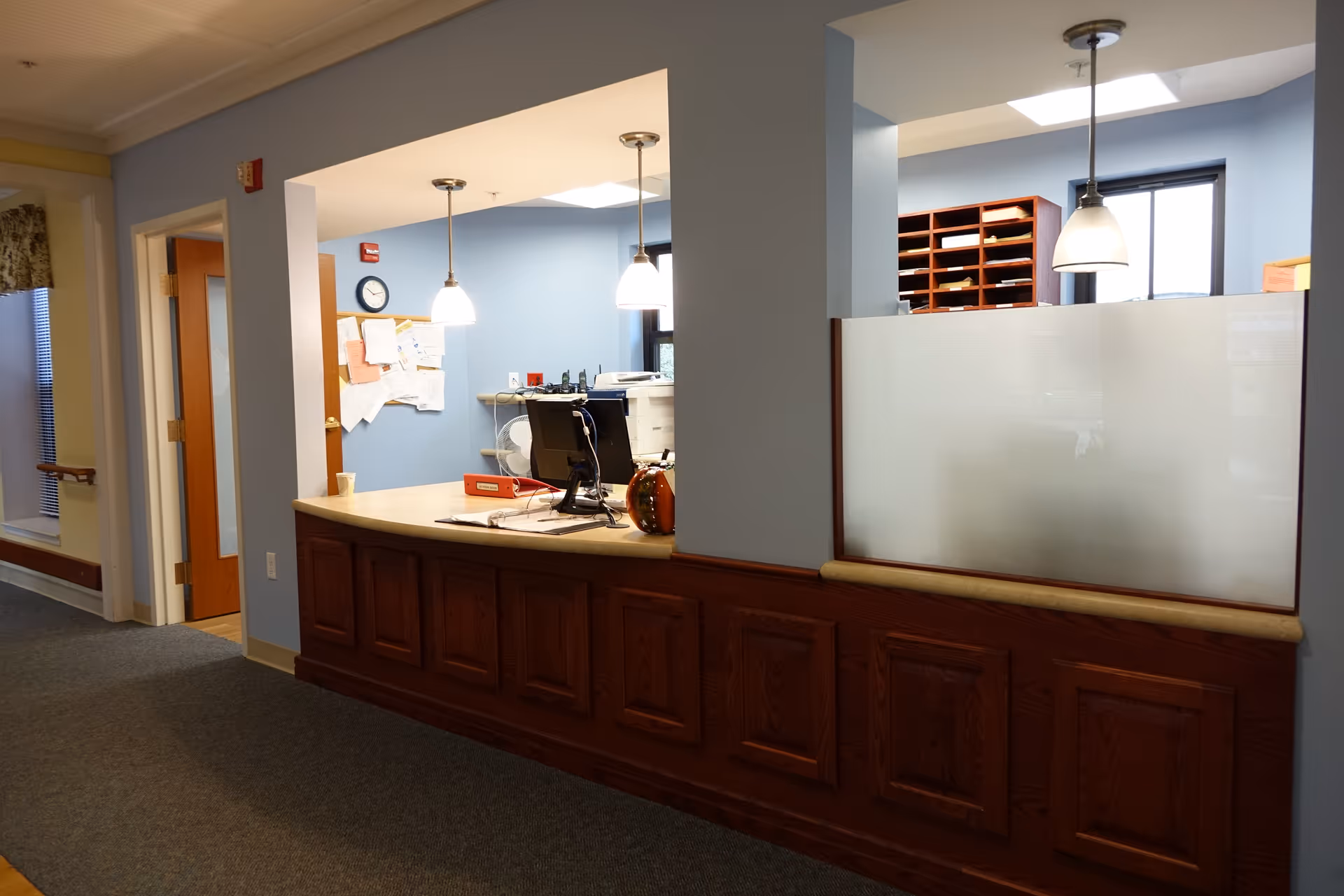 Interior reception desk area with a wooden counter, pendant lights, office equipment, and mail cubbies behind frosted glass.