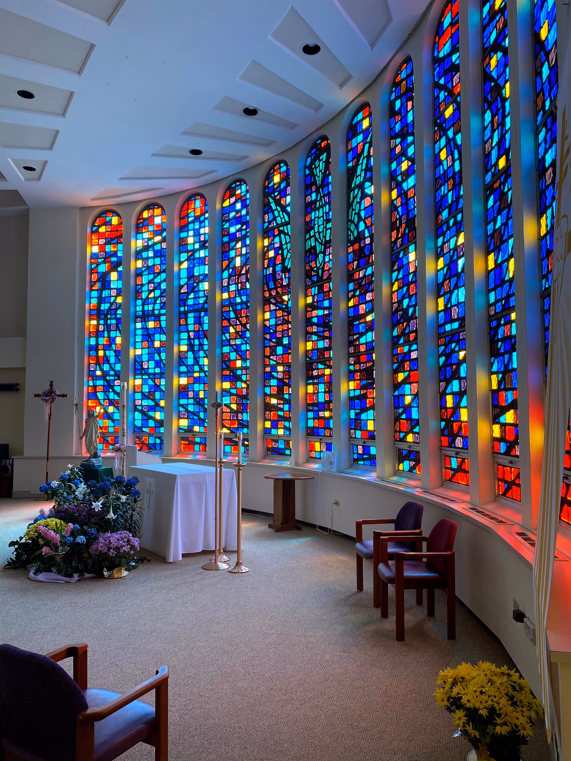 Interior of a chapel or worship room with large colorful stained glass windows along one curved wall. There is an altar covered with a white cloth, a statue, a cross, flower arrangements, and several chairs arranged around the space.