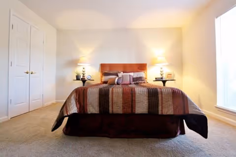 Well-lit bedroom with a centered bed, two nightstands with lamps, closet doors to the left and a window to the right.