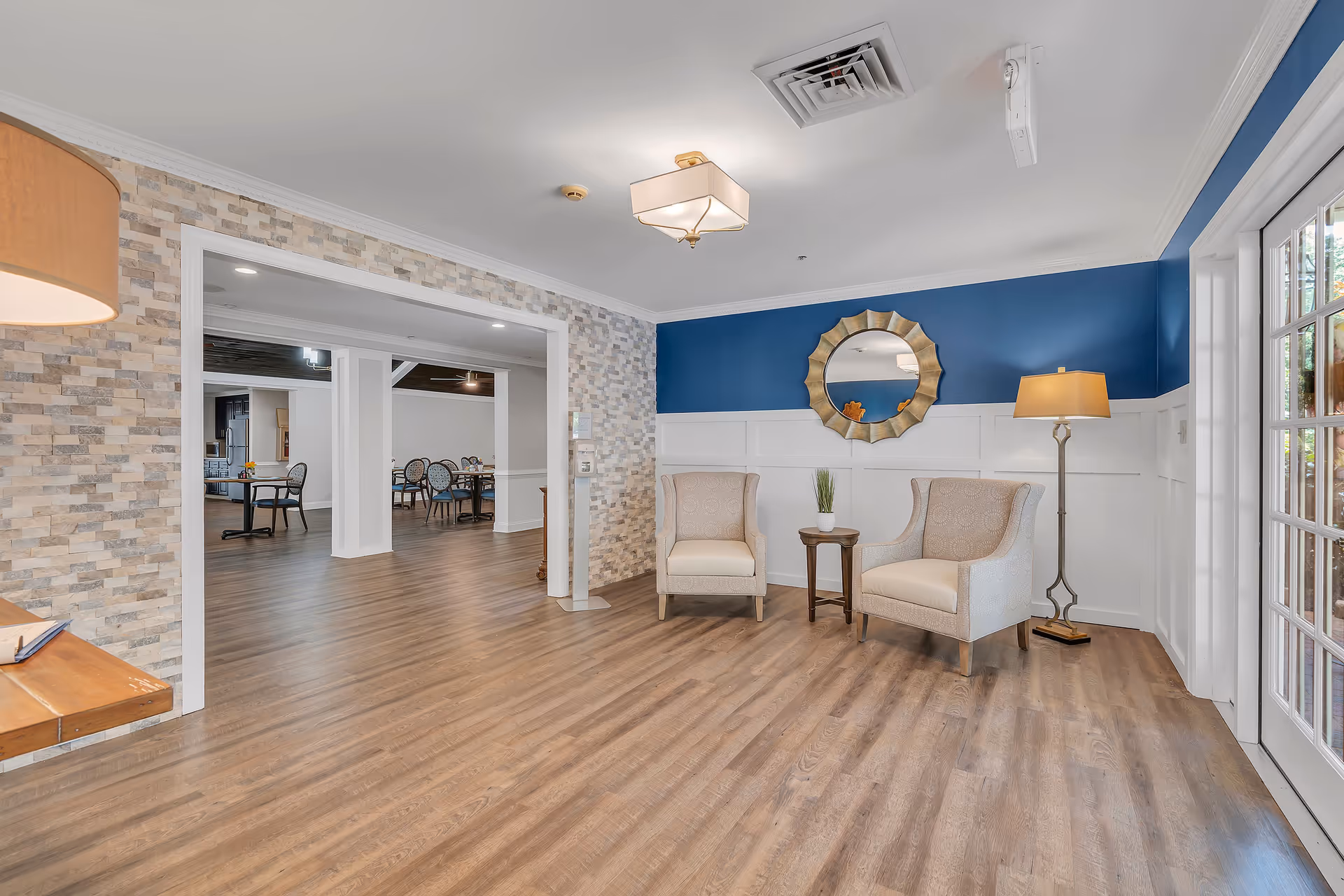 A bright and spacious interior common area with two beige armchairs and a small wooden table between them, placed against a blue and white paneled wall. A decorative round mirror hangs above the chairs. To the right, a tall floor lamp stands near glass doors leading outside. The floor is wood, and the adjacent room visible through a large open doorway contains several dining tables and chairs. The walls near the doorway have a textured stone-like finish.
