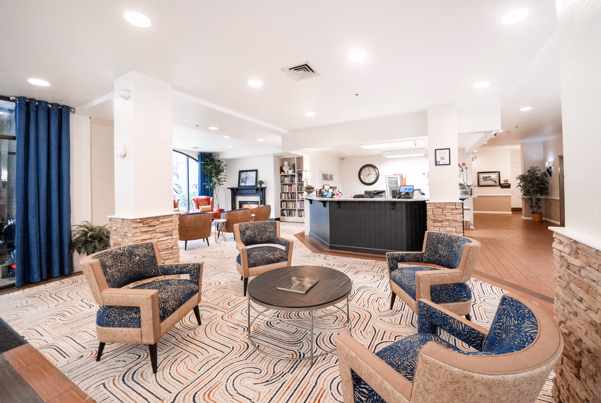 Bright senior living lobby with patterned armchairs around a round table and a reception desk in the background.