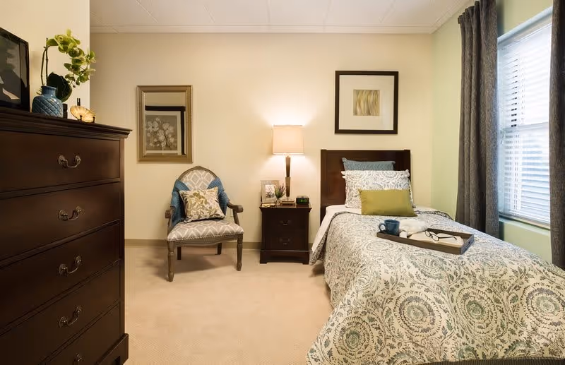 A cozy bedroom with a single bed covered in a patterned bedspread and multiple pillows. A wooden nightstand with a lamp, a framed photo, and a clock sits next to the bed. There is a cushioned armchair with a decorative pillow against the wall, and a tall wooden dresser with a plant and decorative items on top. A window with blinds and dark curtains allows natural light into the room.