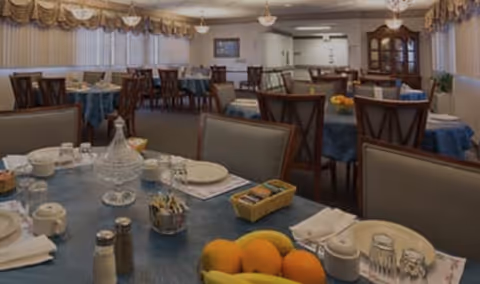 A dining room with multiple tables covered in blue tablecloths, set with plates, cups, and utensils. The room has wooden chairs, a china cabinet, and large windows with blinds and valances. The foreground table has a fruit bowl with bananas and oranges, a glass water pitcher, and various condiments.