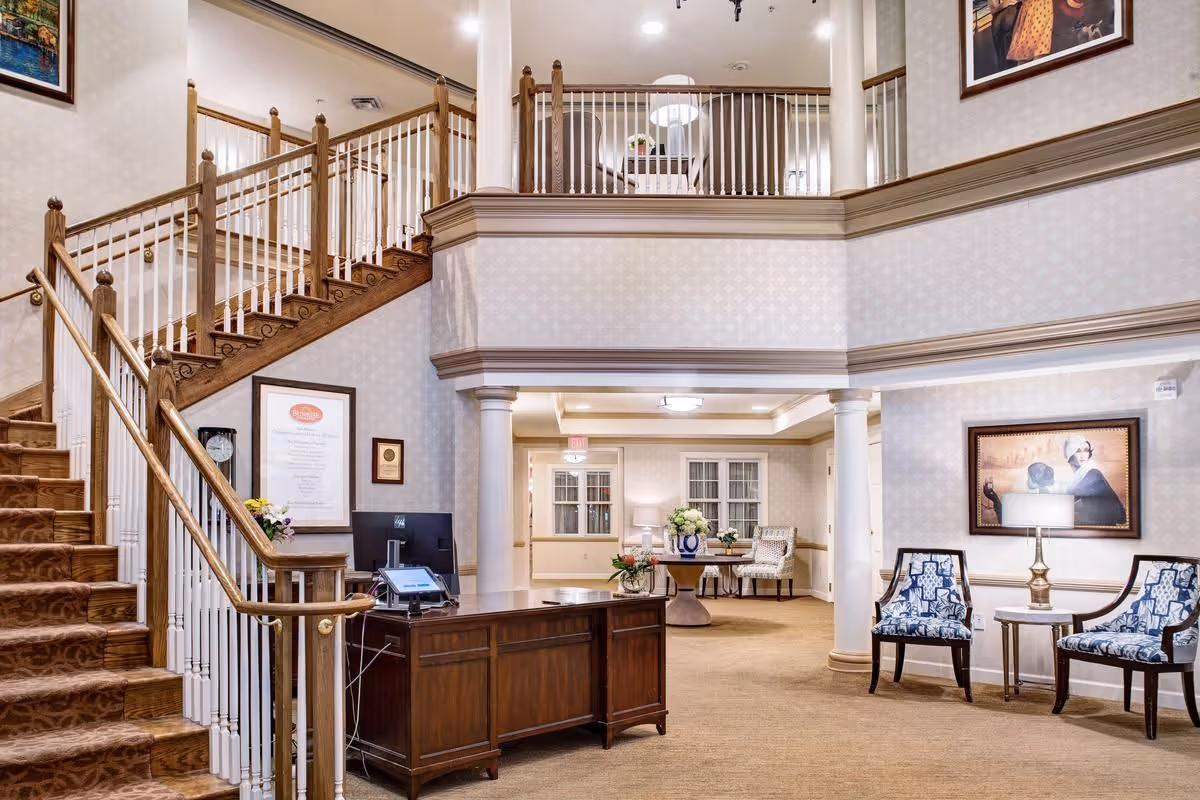 Interior view of a senior living facility lobby with a wooden reception desk, a staircase with wooden handrails and white balusters, two blue patterned chairs with a small round table and lamp between them, and a seating area with a table and chairs in the background. The walls are decorated with framed artwork and the space has a warm, inviting atmosphere.