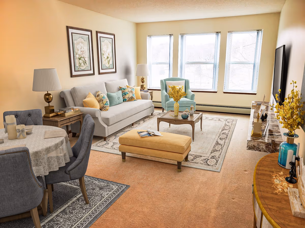 A bright and cozy living room with a beige sofa adorned with colorful pillows, a light blue armchair, and a yellow ottoman on a patterned area rug. There is a wooden coffee table with decorative items and a TV mounted on the wall. A round dining table with a lace tablecloth and four gray upholstered chairs is visible on the left side. The room has large windows letting in natural light, and the walls are decorated with framed floral artwork.