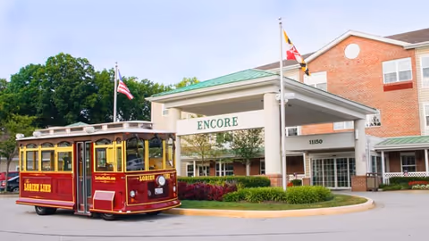 Front entrance of the Lorien Encore senior living facility with a covered porte-cochère, flags, and a red trolley parked at the curb.