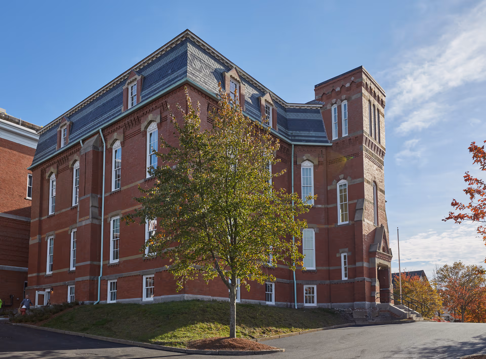 Exterior view of a large, historic red brick building with multiple windows and a slate roof, surrounded by trees with autumn foliage under a blue sky.