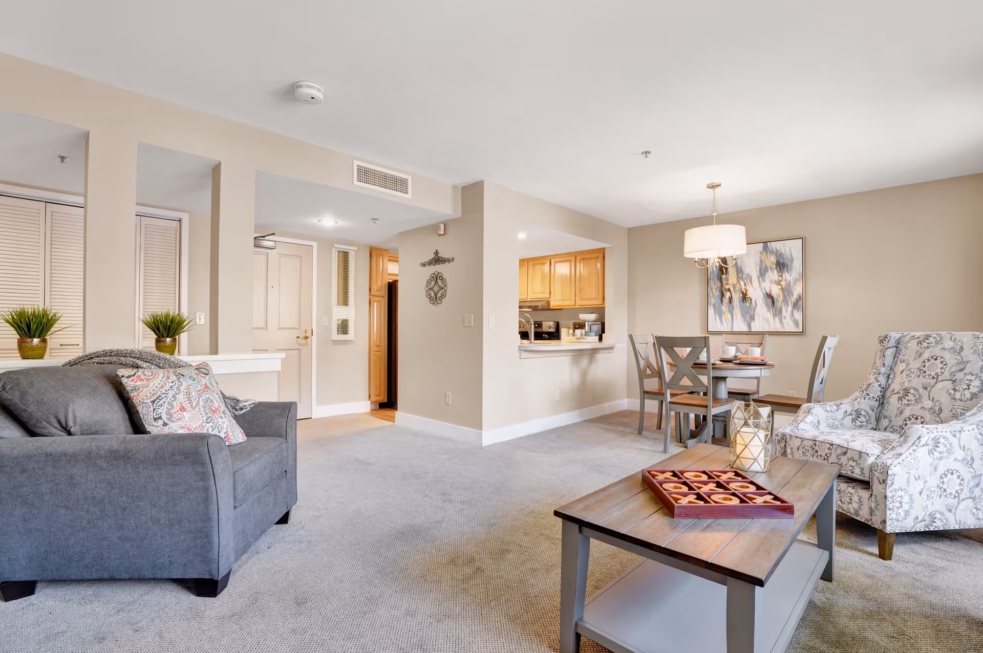 A bright and spacious living room area in a senior living facility with a gray sofa adorned with patterned pillows, a wooden coffee table with a tic-tac-toe game and decorative lantern, a patterned armchair, and a dining table with four chairs. The room has beige walls, carpeted floors, and a kitchen area visible in the background with wooden cabinets.