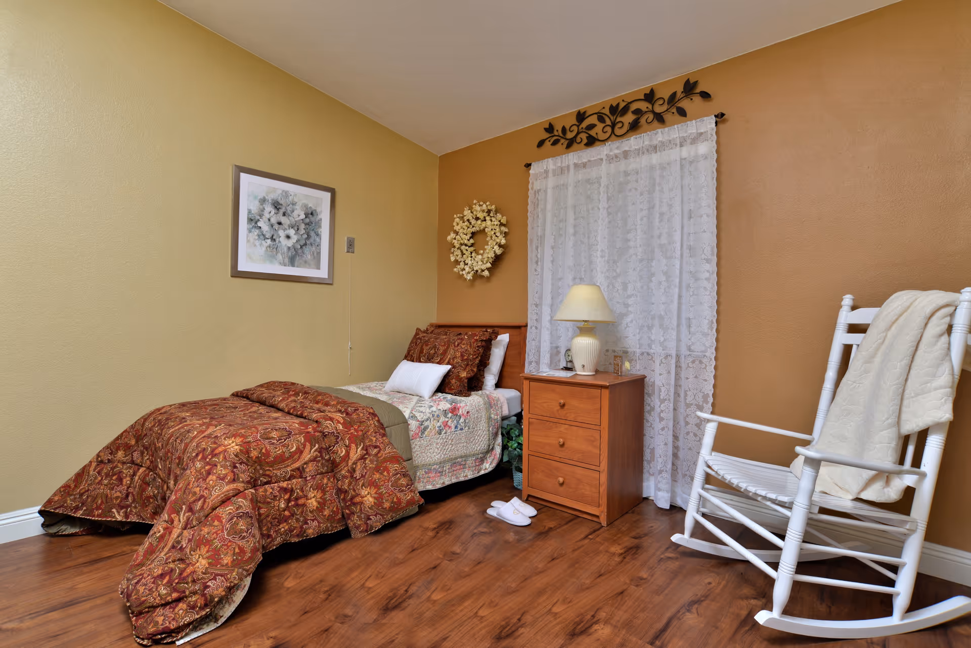 Cozy single bedroom with a twin bed, wooden nightstand and lamp, white rocking chair, and decorative wall accents.