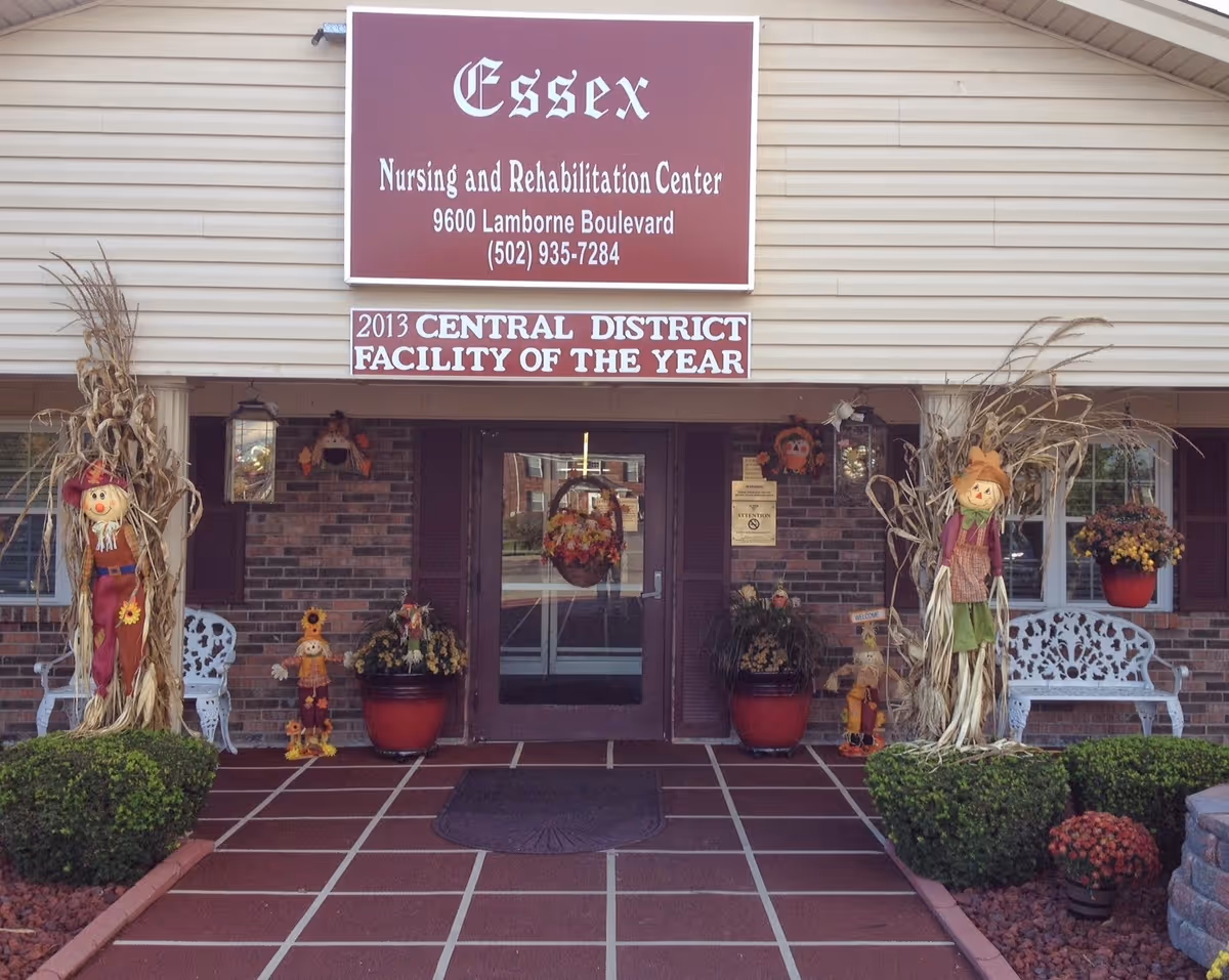 Front entrance of Essex Nursing and Rehabilitation Center with a large sign above the door, fall decorations, potted plants, and benches.