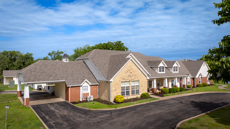 Exterior view of a single-story assisted living facility building with a combination of brick and stone facade, multiple windows, a covered entrance, and a curved driveway surrounded by green lawns and trees under a partly cloudy sky.