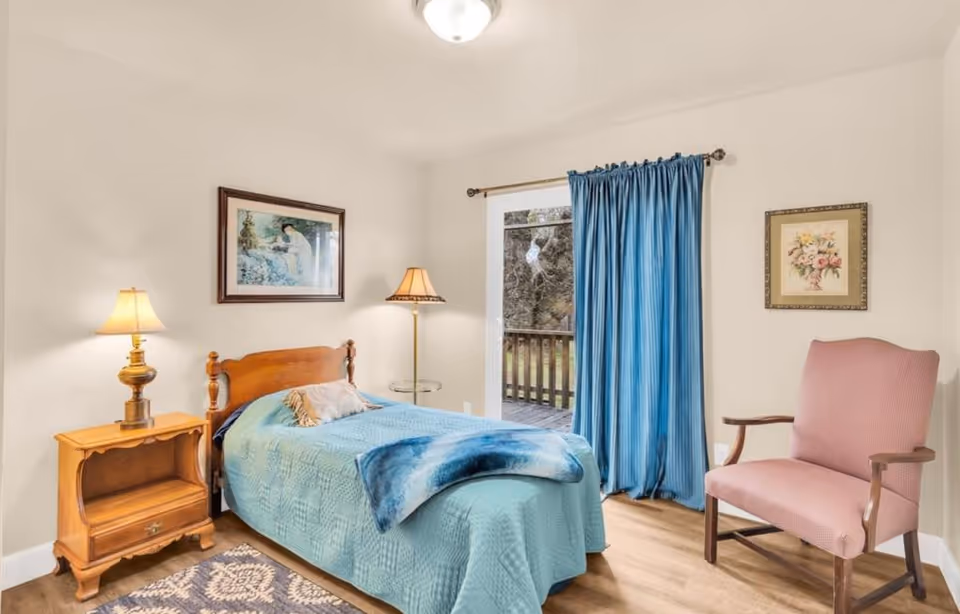 A cozy bedroom with a single wooden bed covered in a light blue quilt and a blue throw blanket. There is a wooden nightstand with a lamp on the left side of the bed and a floor lamp with a glass side table next to it. A pink upholstered armchair is placed on the right side of the room. The room has light-colored walls with two framed pictures hanging, one above the bed and one above the chair. Blue curtains cover a glass door that leads to an outdoor wooden deck.