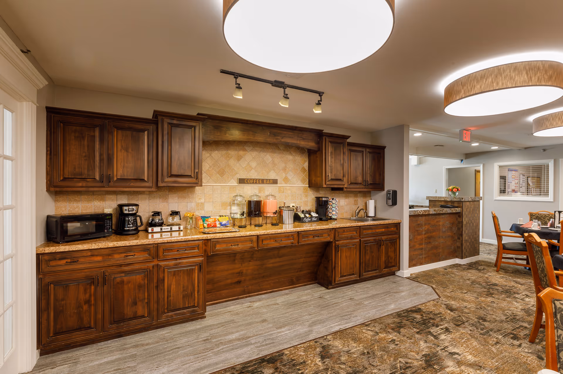 Interior view of a coffee bar area in a senior living facility with wooden cabinets, a microwave, coffee maker, beverage dispensers, and snacks on the counter. Adjacent to the coffee bar is a dining area with tables and chairs, illuminated by large circular ceiling lights.